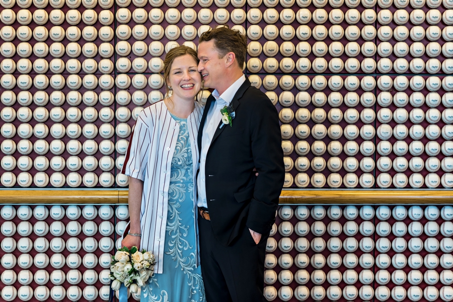 A newly married couple hold each other in front of a wall of baseballs at a Phillies game.